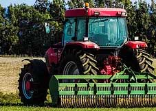 red combine harvester drives over a green field on a farm with trees in the background and a blue sky above