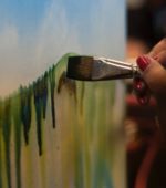 paintbrush moving across canvas adding green details under a painted sky
