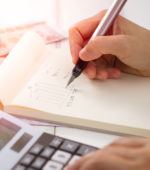 hands hold a pen to a notepad with another hand punches numbers on a calculator