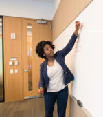 professor writes on white board in front of college class