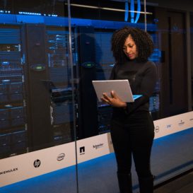 woman in suit stands holding a laptop outside of a blue-lit server room