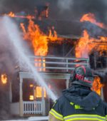 fire fighter stands in front of burning building holding hose spraying water