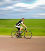 bicyclist riding on a road against a grassy hill and a blue sky dotted with clouds